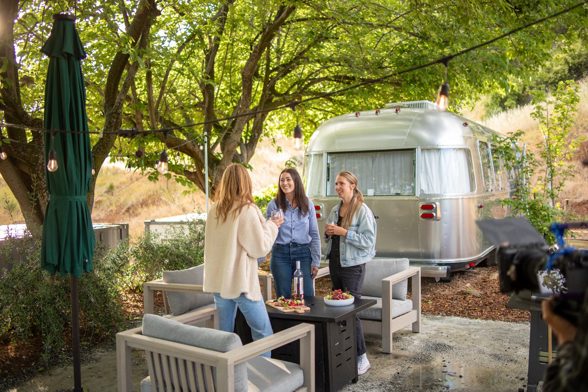 Guests sharing drinks around a fire table beside an Airstream under oak trees