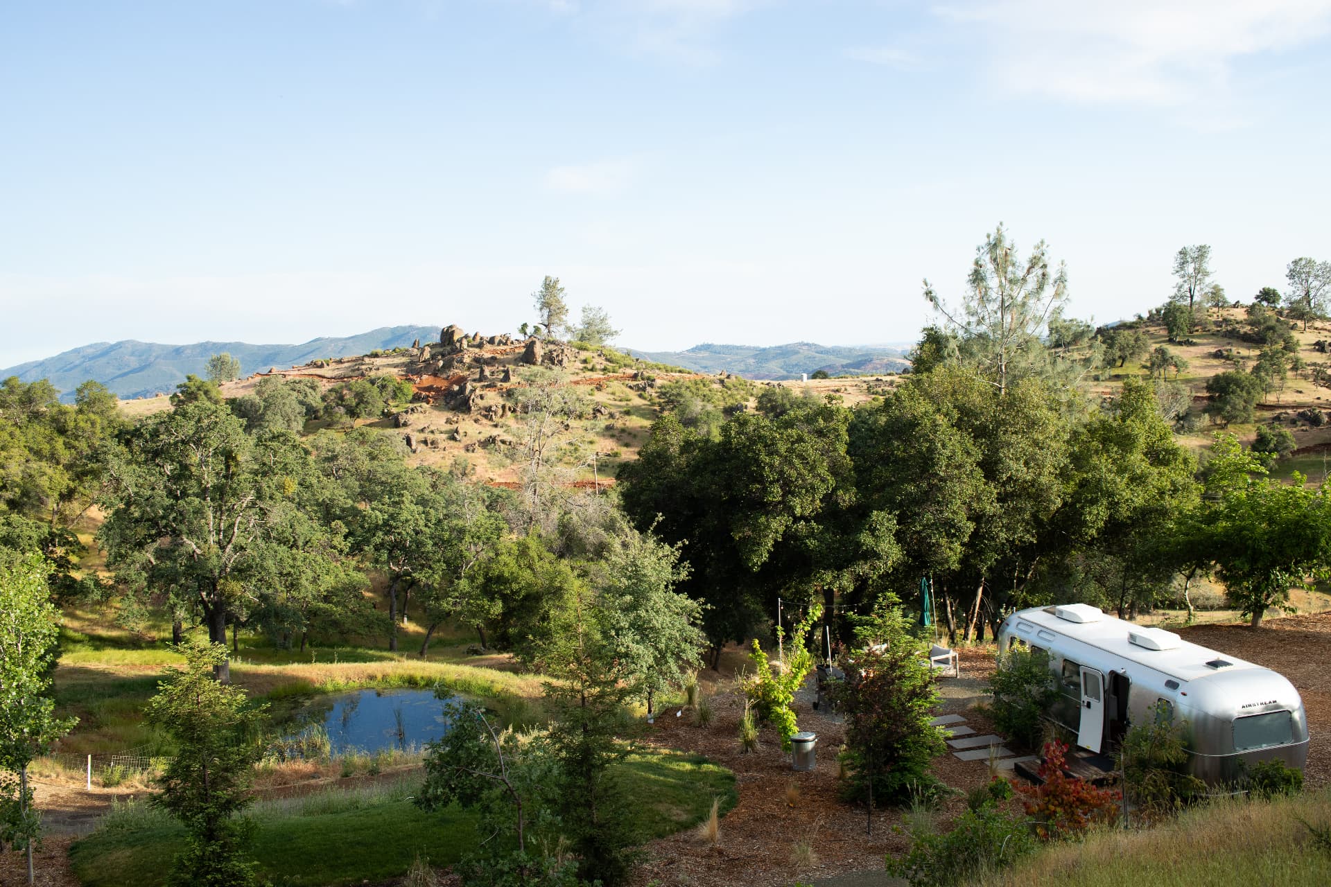 Wide view over the property's oak-studded hills, a pond and an Airstream below distant mountains